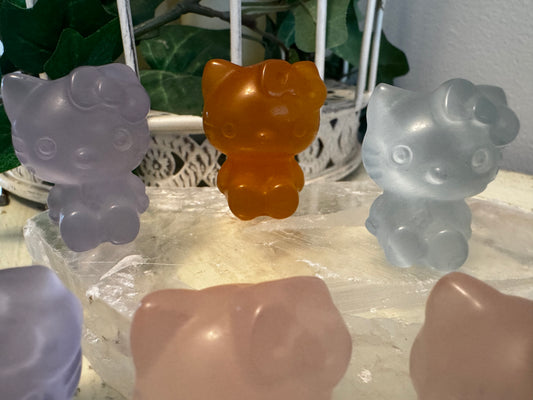 Three Hello Kitty figurines made of translucent orange, blue, and light lavender stone, sitting on a rock slab.