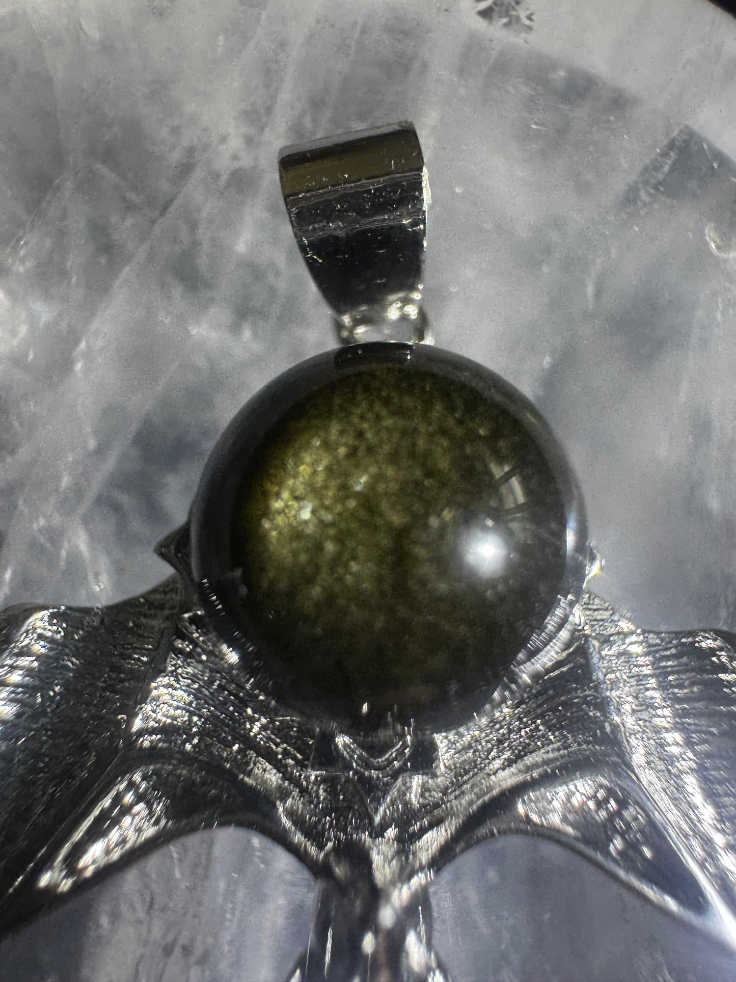 Close-up of a gold sheen obsidian sphere pendant with a silver bail and setting against a light gray textured background.