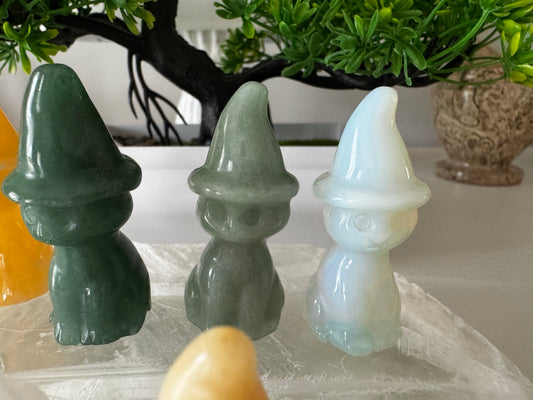 Three crystal gnome figurines in green, grey, and opal white, sitting on a crystal slab in front of a decorative plant.