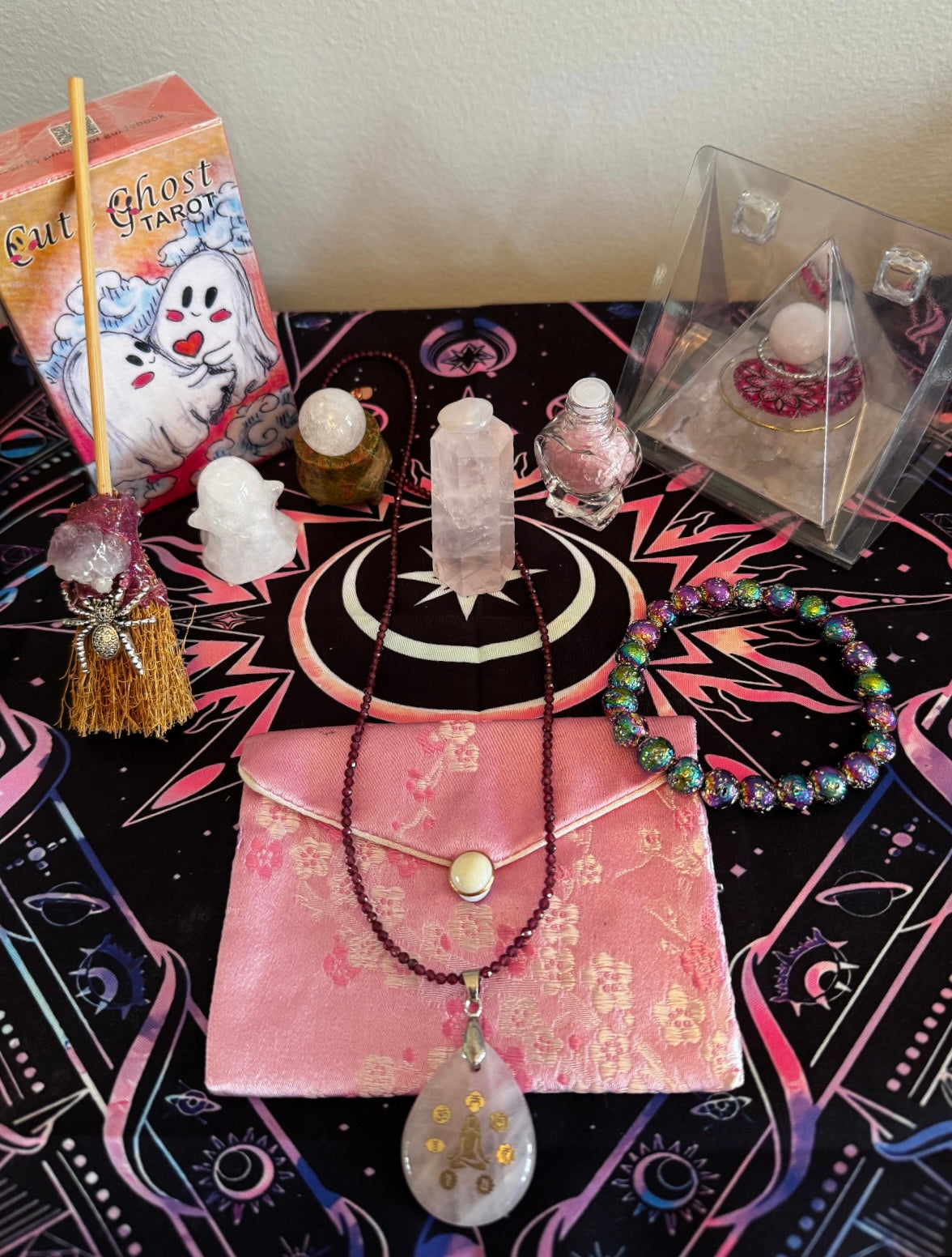 Product display featuring a tarot deck, crystals, jewelry, and other spiritual items arranged on a patterned tablecloth.