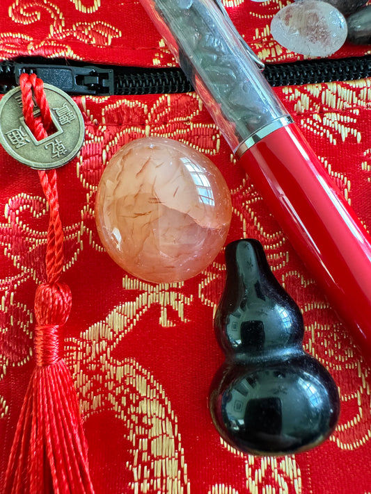 Still life of a red and gold embroidered pouch with a zipper, adorned with a coin tassel, an orange sphere, a black gourd, and a red pen.