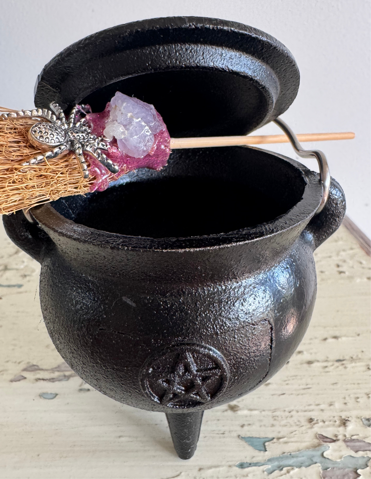 Close-up of a black, three-legged cauldron with a lid, adorned with a small broom featuring a silver spider and a purple crystal.