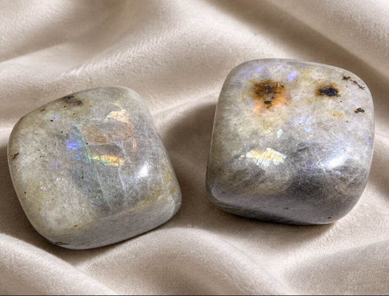 Two polished tumbled labradorite stones with a milky white and gray base and iridescent rainbow flashes, resting on a light surface.