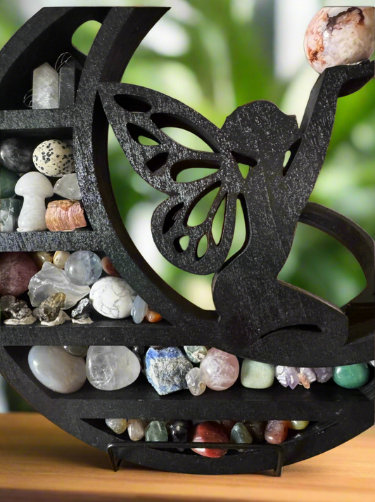 Close-up of a crescent-shaped shelf filled with rocks and crystals. A black silhouette of a fairy is inlaid on the front, and the shelf is set on a wooden surface. The background is blurred green foliage.
