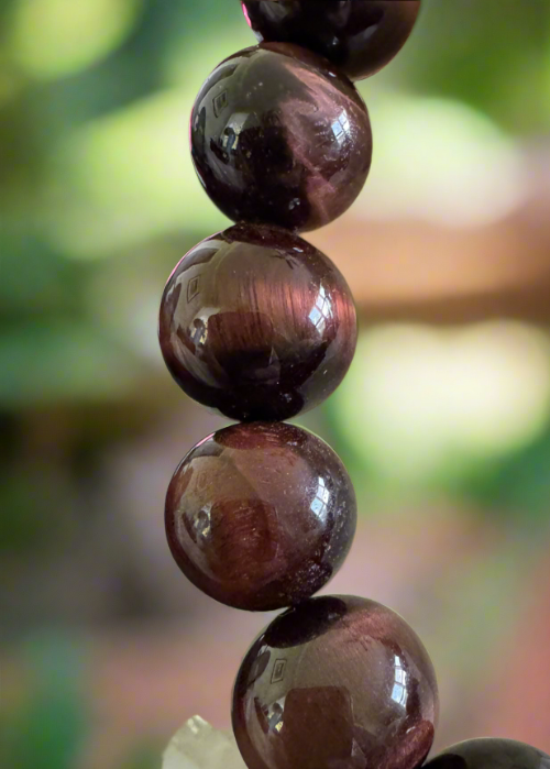 Close-up of a burgundy gemstone bead bracelet with reflections, against a blurred green and brown background.