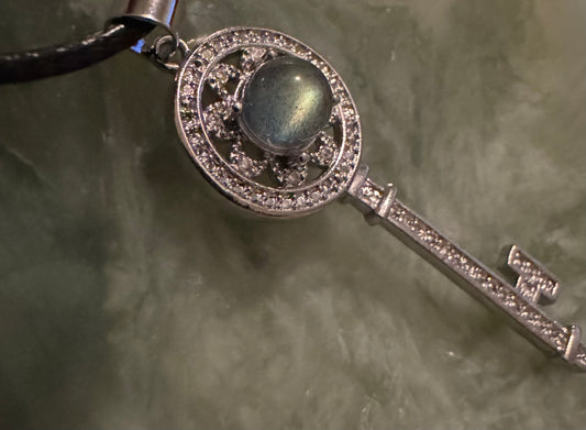 Close-up of a silver key pendant with a round blue gem, adorned with rhinestones, hanging from a black cord on a muted green surface.