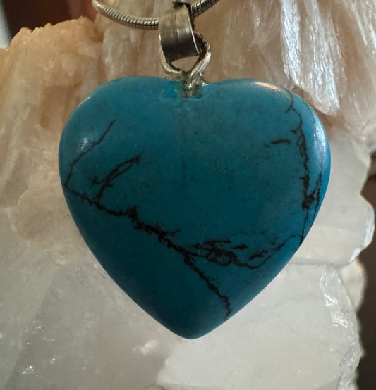 A turquoise heart-shaped pendant with dark veining hangs from a silver chain against a white crystalline background.