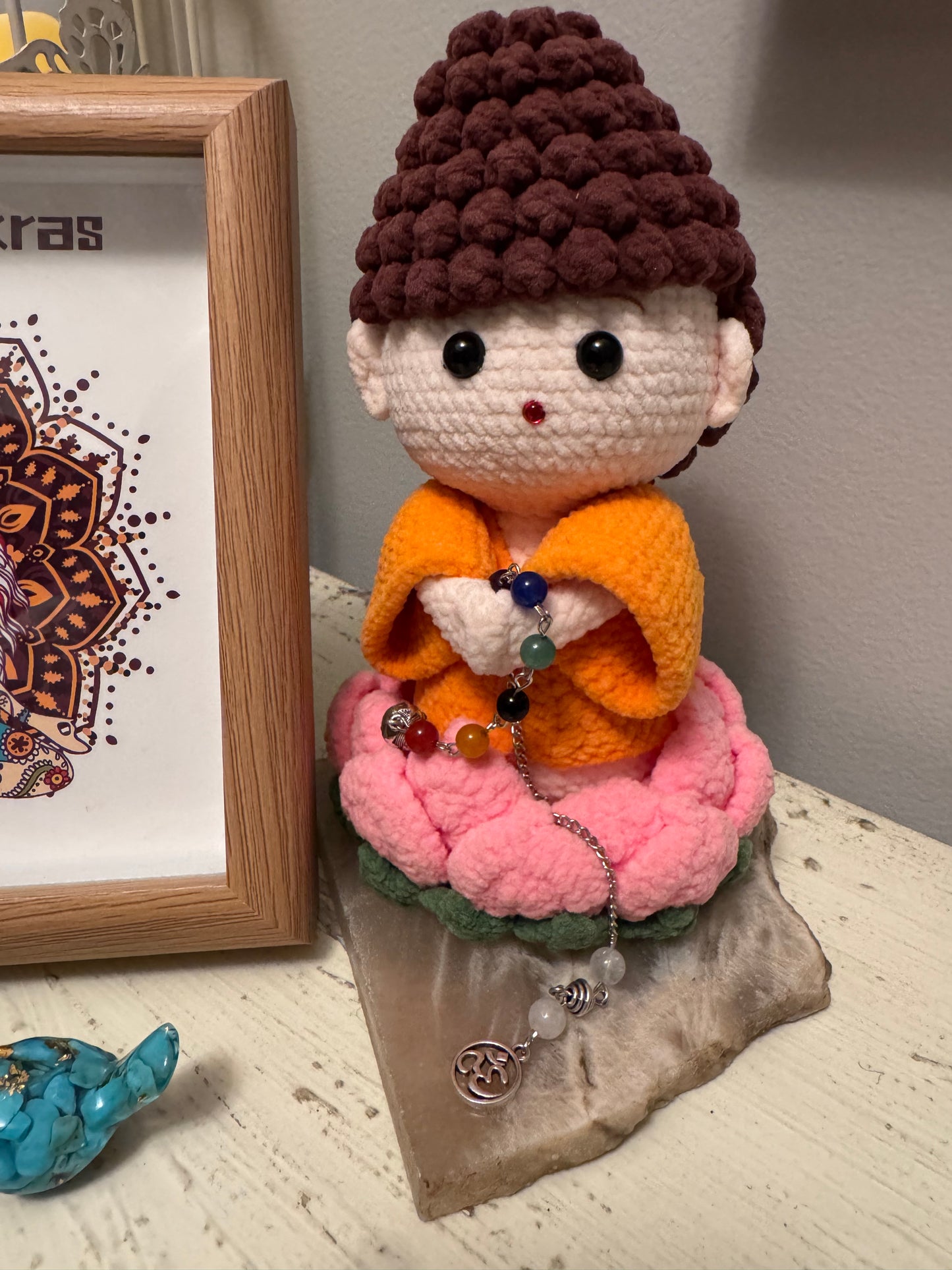 Crochet Buddha doll in orange robes, seated on a pink lotus flower, holding a chakra bead chain.