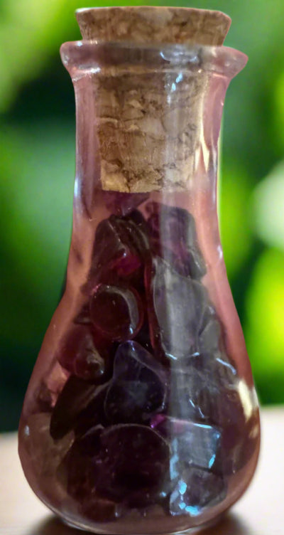 Small, pear-shaped pink glass bottle with cork stopper and amethyst stones inside. Green, blurred background.