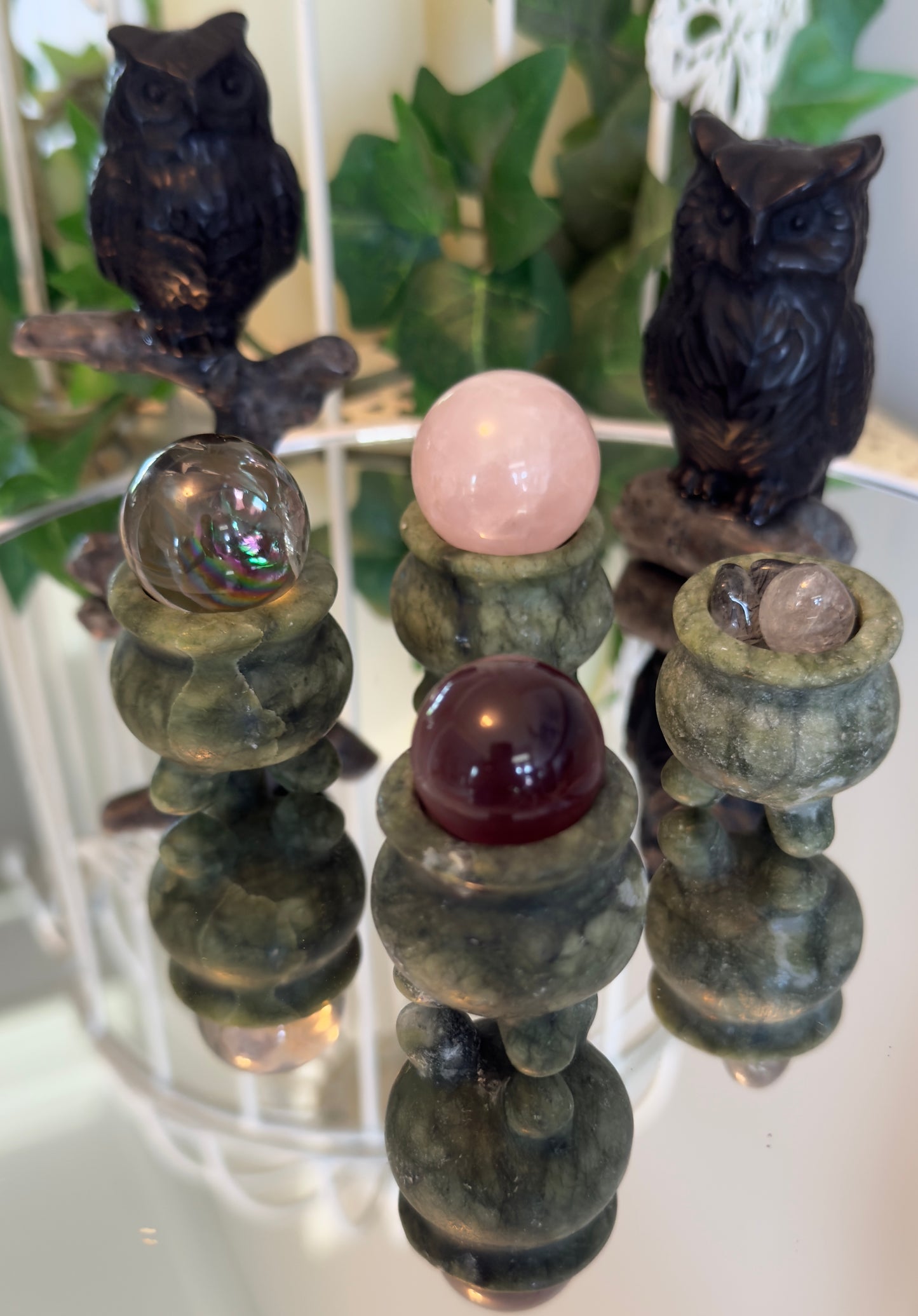 Two owl figurines sit on a reflective surface, behind three crystal balls on ornamental jade stands.