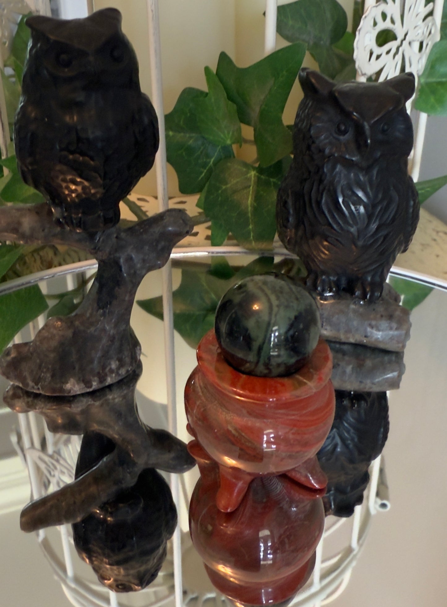 Two carved stone owls and a ball-and-base sculpture on a mirrored surface in front of greenery and a white decorative object.