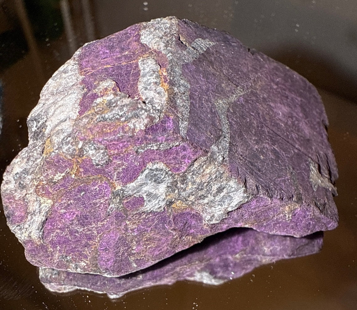 Purplish-gray mineral specimen, possibly sugilite, resting on reflective surface.