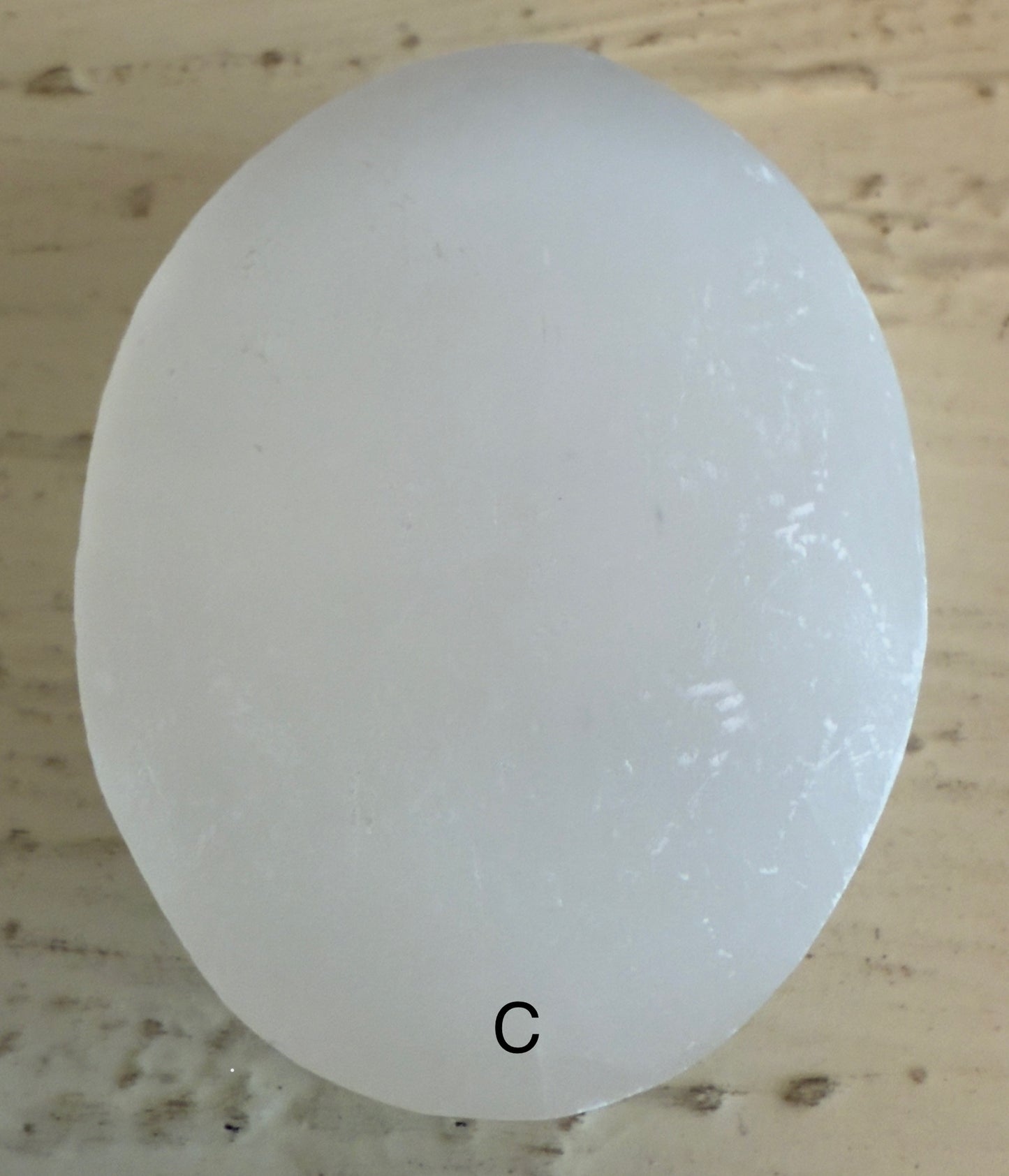 Oval, translucent white stone with the letter "C" at the bottom on a textured tan surface.