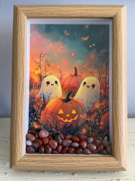 A framed artwork with a Halloween theme, featuring two smiling ghosts, pumpkins, and decorative stones.