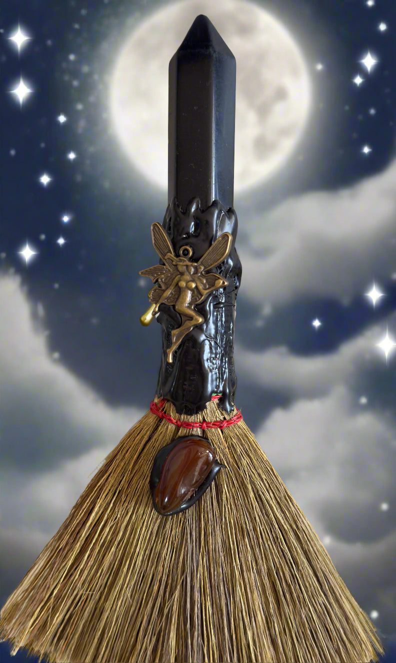 A witch's broom with a black crystal on top, adorned with a brass fairy and a brown stone against a night sky backdrop.