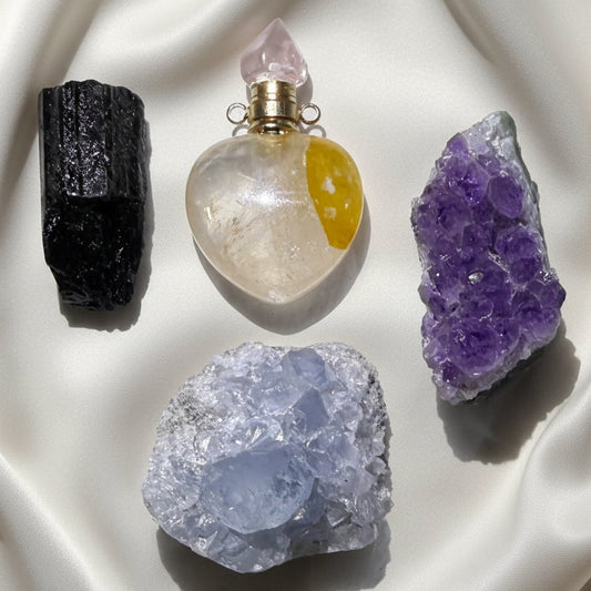 Four raw crystals and a heart-shaped glass perfume bottle arranged on a white textured background. Black Tourmaline, Golden Healer Heart, Amethyst Geode and Celestite Geode