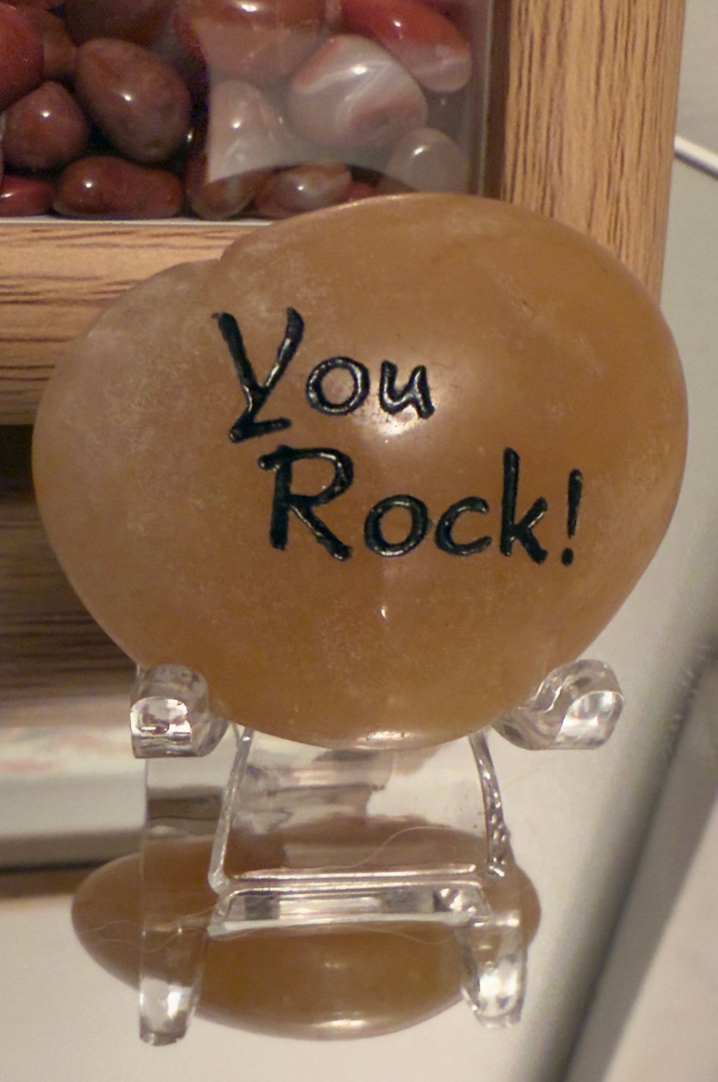 Orange Calcite Inspiration Stone with the engraved phrase “You Rock!” displayed on a clear stand, all placed on a reflective surface. Main Street Crystals and Minerals