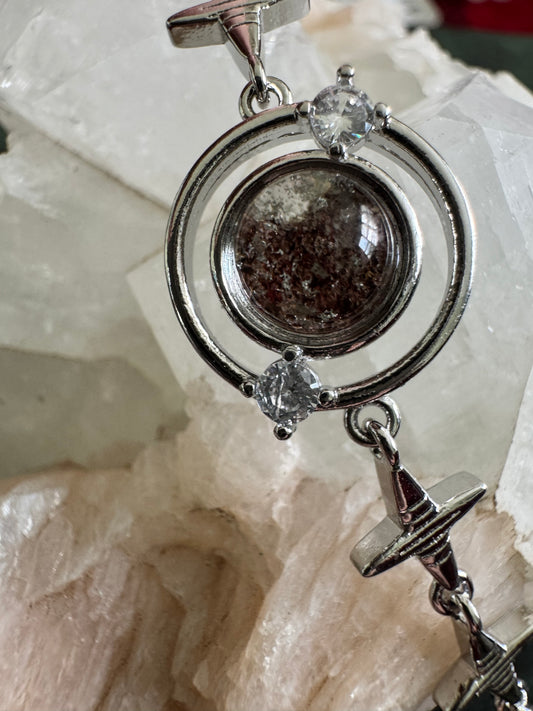 Silver pendant featuring a double-ring design with clear stones and a central, speckled dark stone. A cross charm hangs below. The pendant is resting on a light-colored, textured mineral or stone surface.