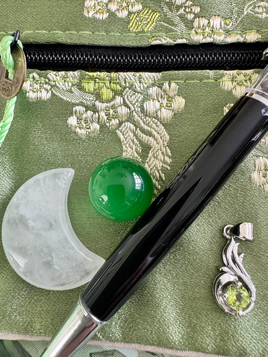 Close-up of a green patterned fabric pouch with white crescent moon stone, green jade ball, black and silver pen, and a silver peridot pendant.