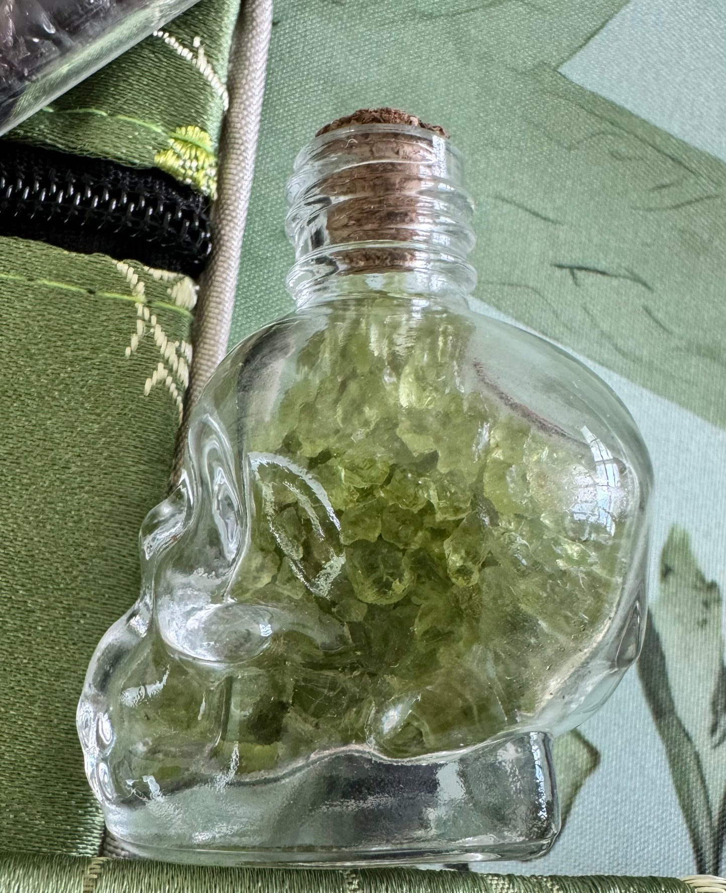 Skull-shaped glass bottle filled with green crystals, sealed with a cork stopper.