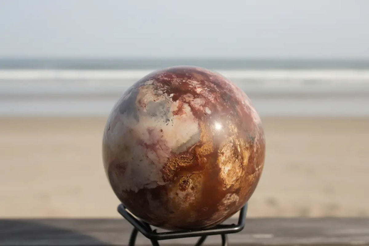 Polychrome jasper sphere in a small stand on the beach Colors are bright oranges, blue, ivory and white