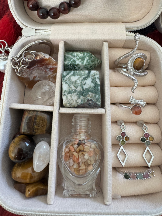 A jewelry box containing an assortment of gemstones and jewelry including a tiger's eye snake ring, tourmaline earrings and bracelet, and tumbled stones.