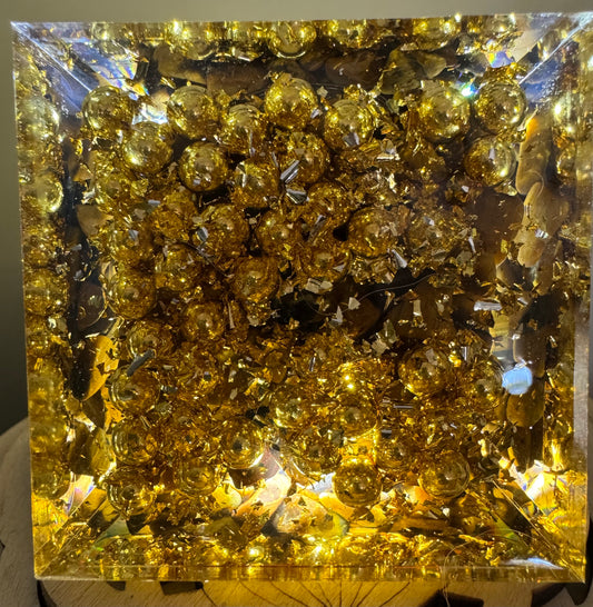 Square, clear resin block with embedded golden spheres, gold leaf flakes, and quartz pieces.