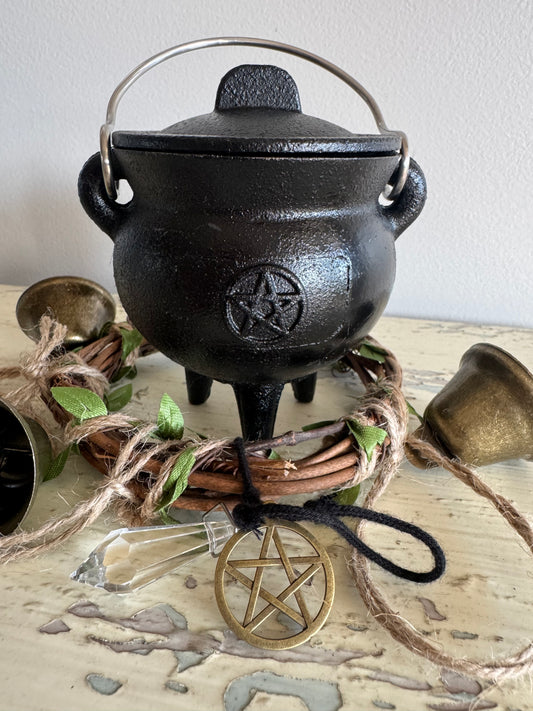 Triple moon pentacle black cast iron cauldron with lid, three legs, and metal handle, surrounded by rustic altar decor, crystal point, and gold pentagram ornament—perfect for witchcraft rituals, spellwork, and Wiccan altar supplies