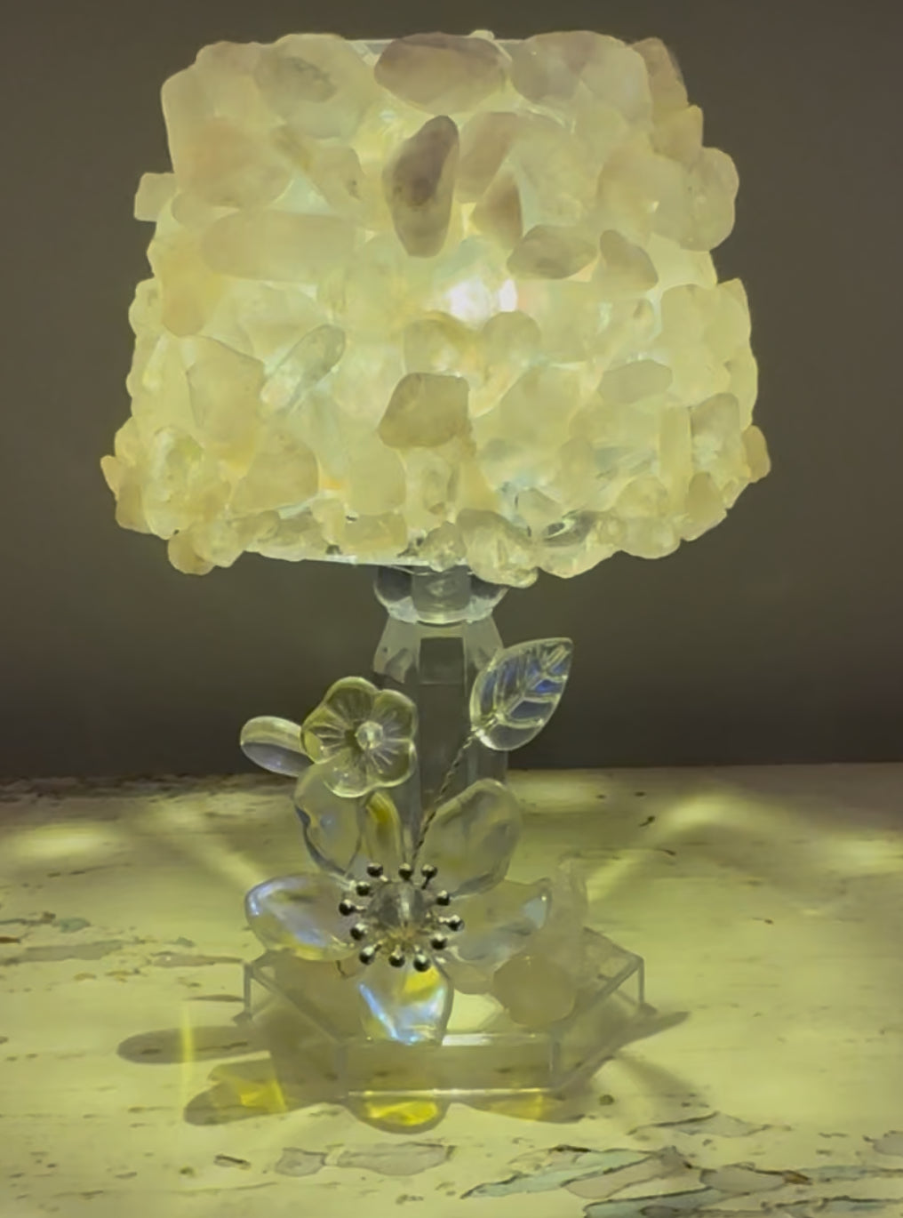 Illuminated lamp featuring a shade covered in translucent, yellowish stones, atop a clear, floral-themed base.