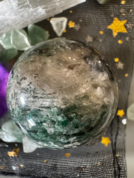 Close-up of a crystal ball with green and gray inclusions, on a star-spangled gray cloth, surrounded by other crystals.