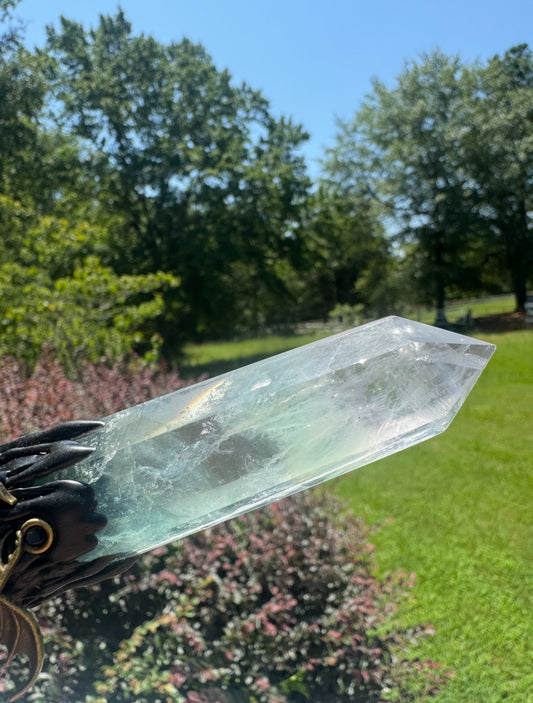A large clear crystal point, held in a dark, ornate holder, with green trees and grass in the background.