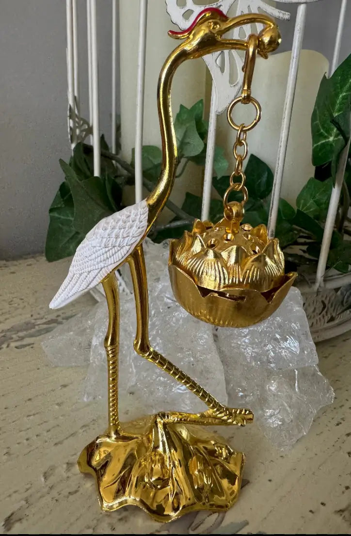 A gold-toned decorative crane figurine holding a lotus-shaped bowl, with a white textured wing and a small red crest on the head, displayed on a light surface with clear crystal and green leaves in the background.