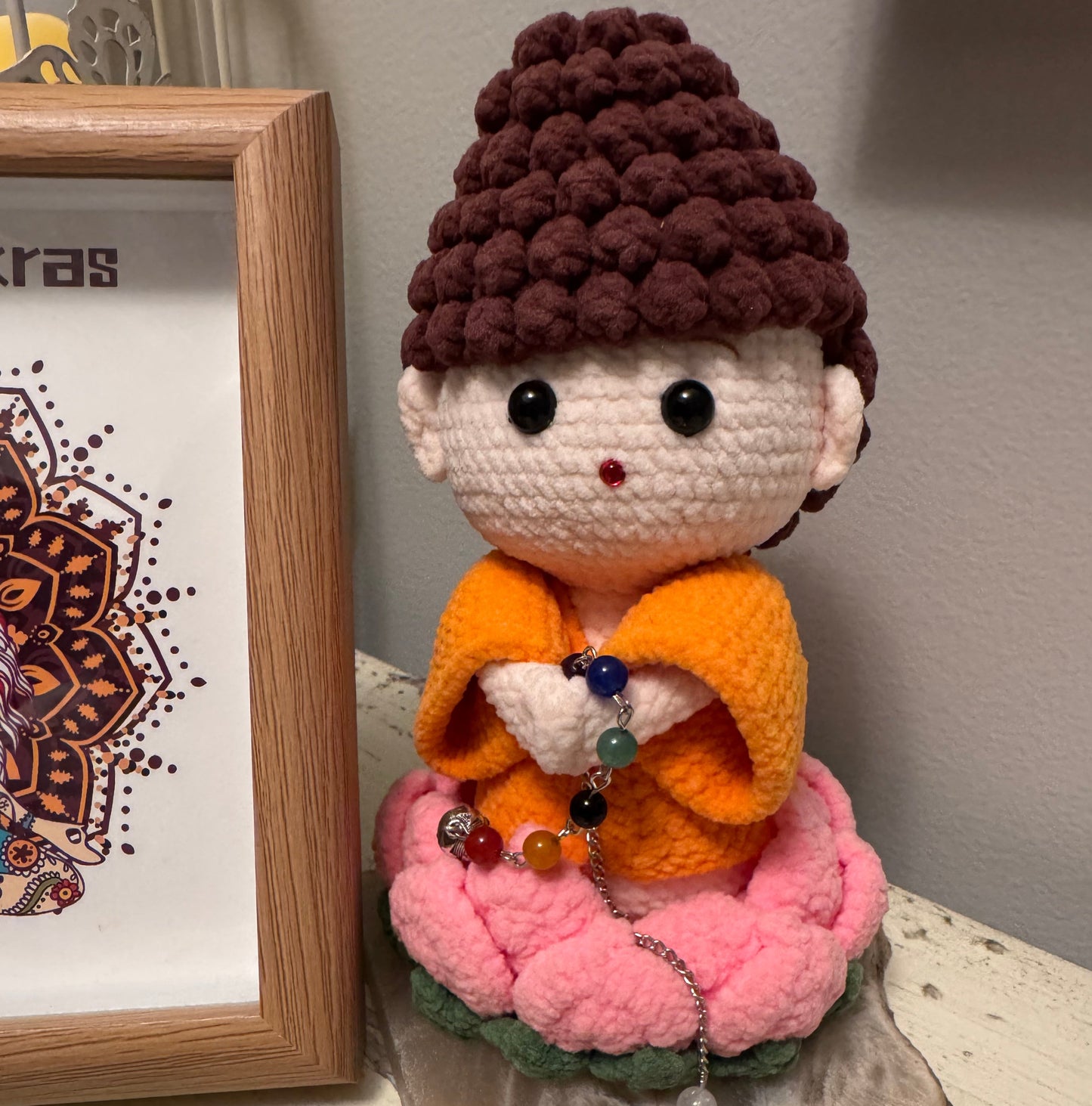 Crochet Buddha doll in orange robes, seated on a pink lotus flower, holding a chakra bead chain.