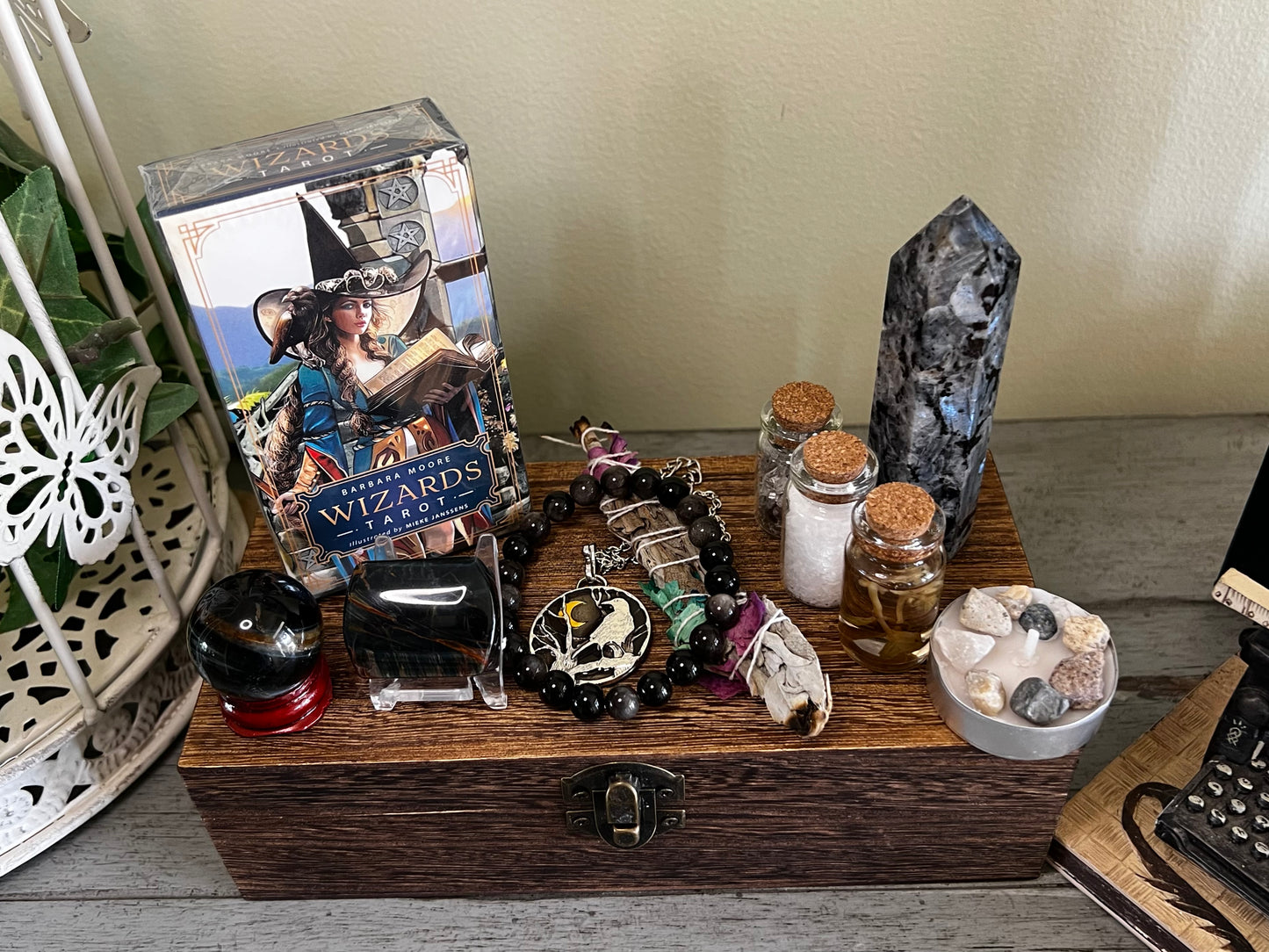 Still life featuring the Barbara Moore Wizards Tarot deck, crystals, sage, and vials arranged on a wooden box with a metal latch. The tarot deck depicts a woman in a hat and blue jacket reading a book. Other items include a black crystal sphere on a red base, a tower crystal, and a beaded necklace with a raven pendant. A miniature typewriter sits to the right.