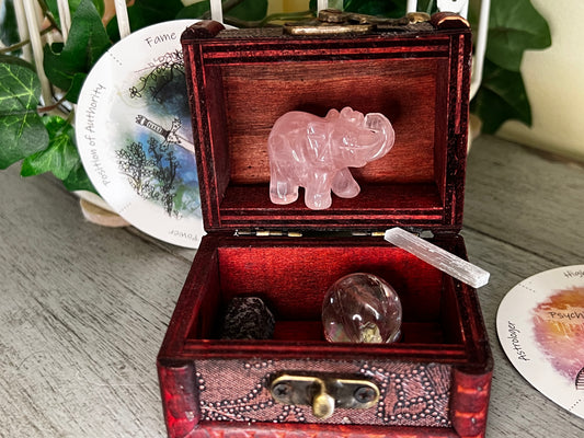 An open, decorative wooden box filled with a rose quartz elephant carving, a selenite wand, and two stones, sitting next to oracle cards and greenery on a wooden surface.