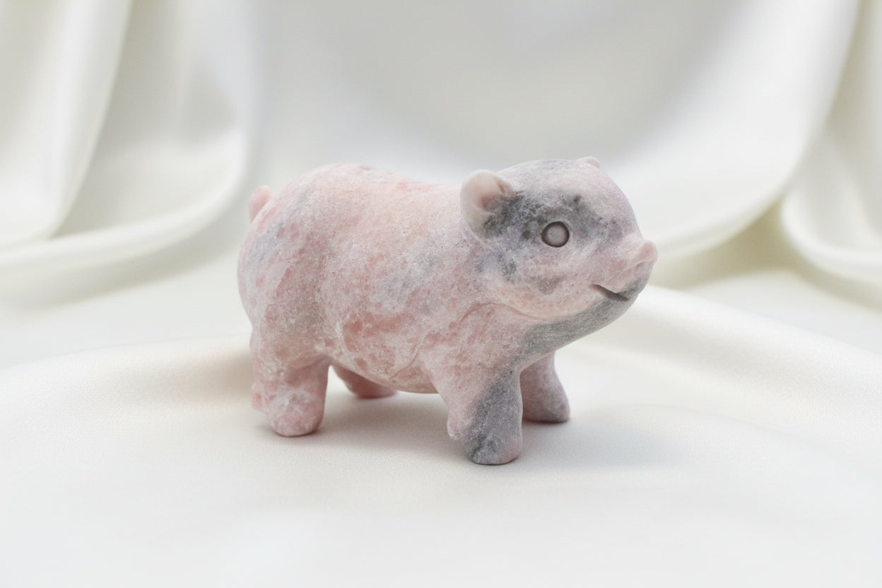 A small pig figurine carved from pale pink calcite stone, standing on a tabletop with other decorative objects blurred in the background.