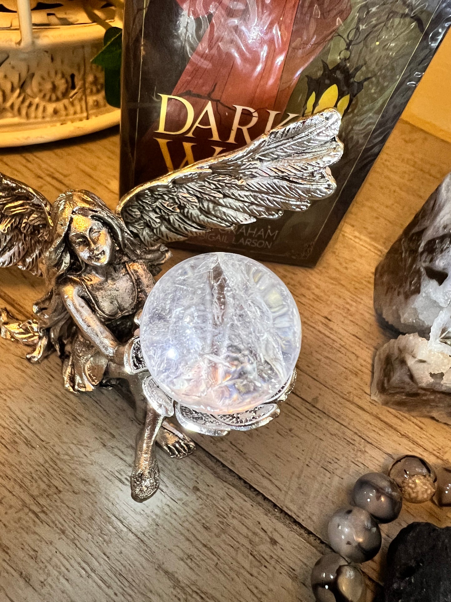 Silver angel figurine holding a crystal ball, with a book and pebbles on a wooden surface.