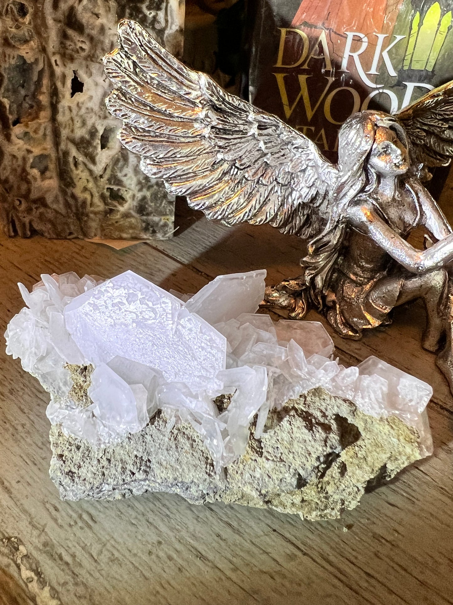 Close-up of a quartz crystal cluster on a rough, earthy matrix, next to a silver angel figurine, on a wooden surface. A book titled "Dark Wood" is in the background.