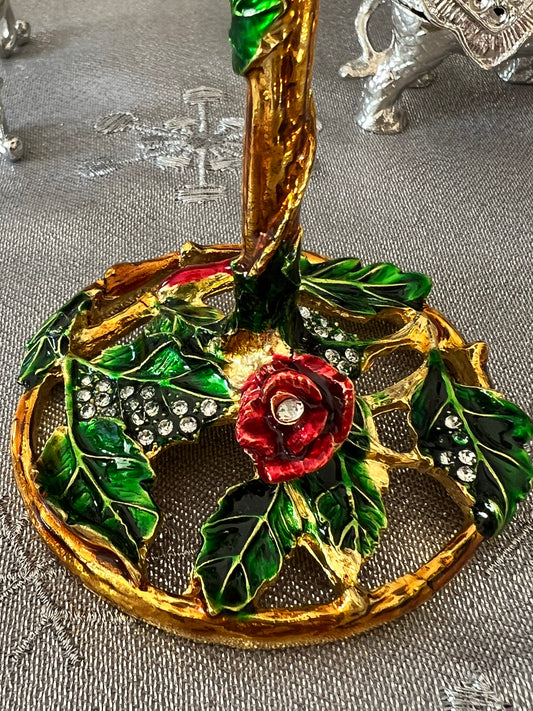 Close-up of a gold and enamel decorative object with a red rose and green leaves, accented with clear stones.