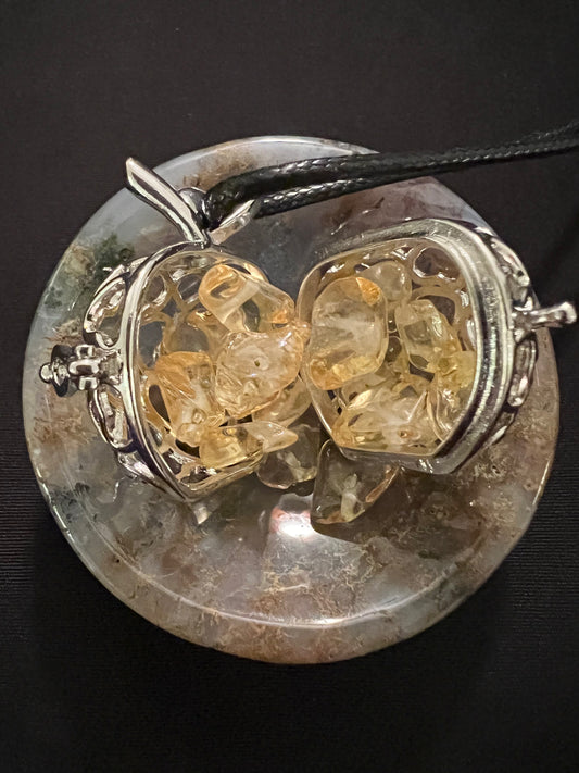 An open, heart-shaped, silver pendant filled with yellow gemstone chips, on a patterned stone, set against a black background.