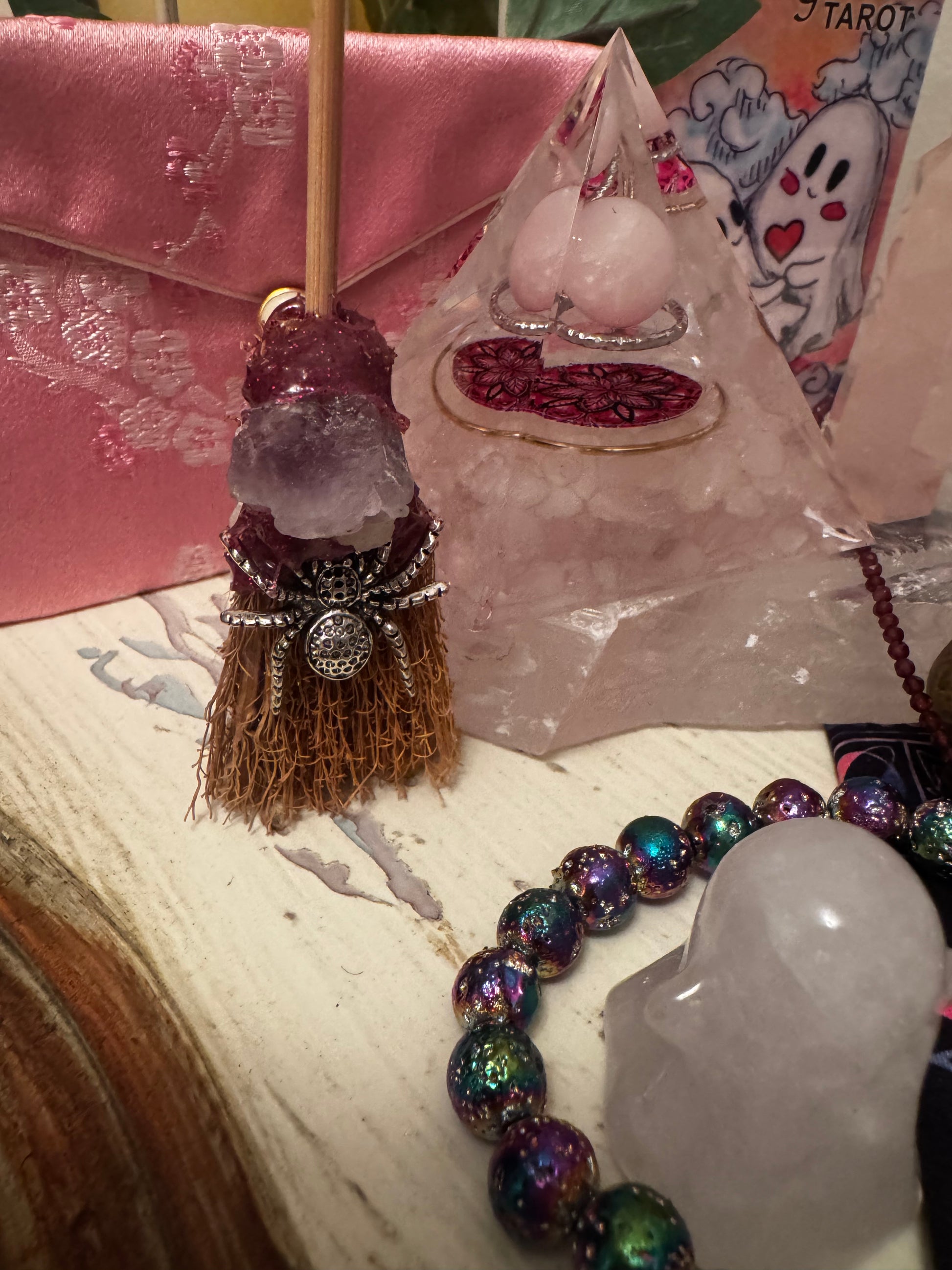 Close-up of a witchy, eclectic collection, including a small crystal broom with a silver spider, a pink satin pouch, a crystal pyramid with jewelry inside, a ghost tarot card, a bead bracelet, and a crystal ghost.
