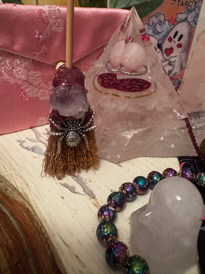 Close-up of a witchy, eclectic collection, including a small crystal broom with a silver spider, a pink satin pouch, a crystal pyramid with jewelry inside, a ghost tarot card, a bead bracelet, and a crystal ghost.