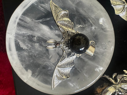 Overhead shot of a silver bat pendant with a black stone on a clear round dish.