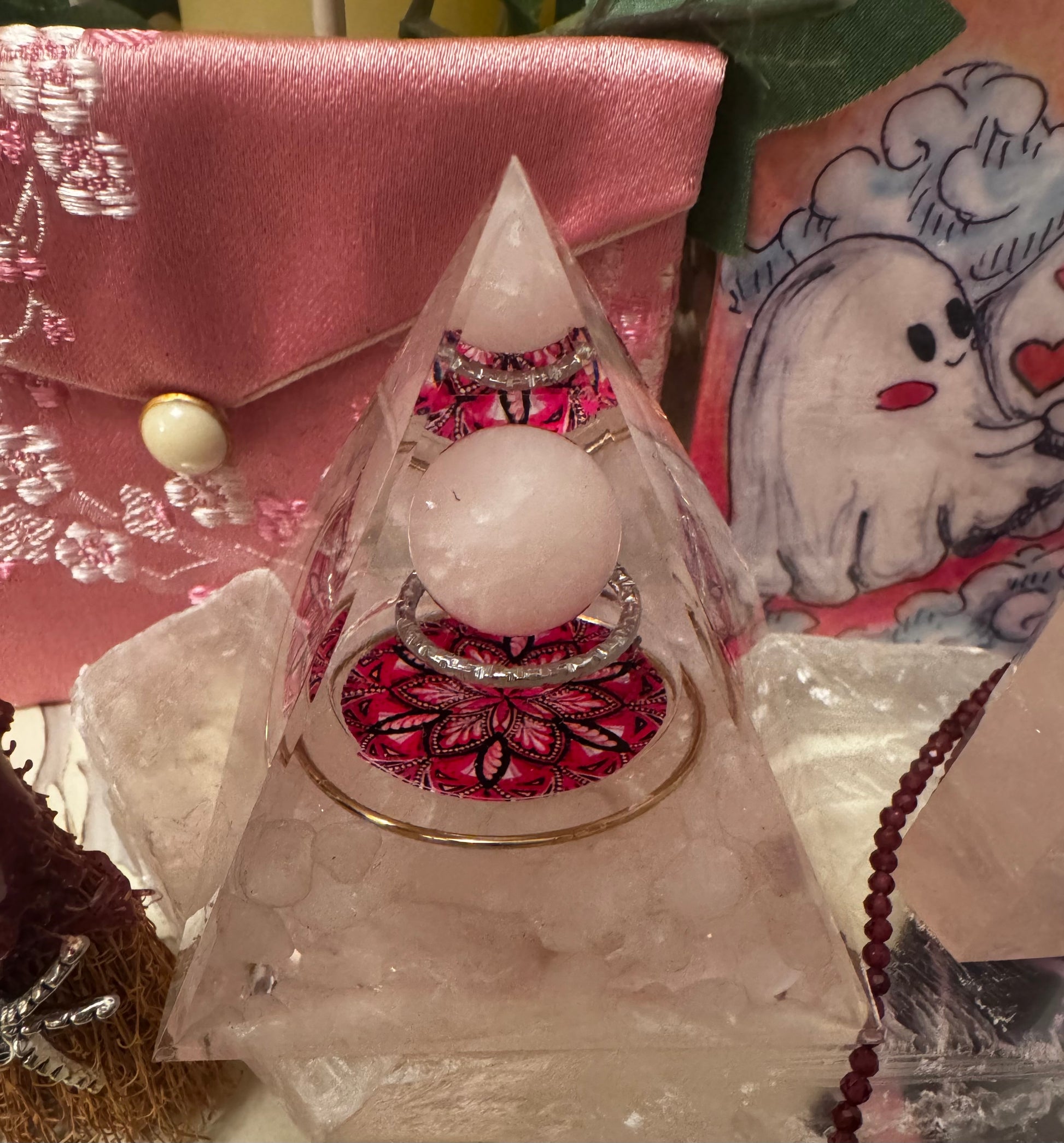 Close-up shot of a pink and white display, featuring a pyramidal crystal sculpture encasing jewelry, a floral pink pouch, and a cartoon ghost illustration.