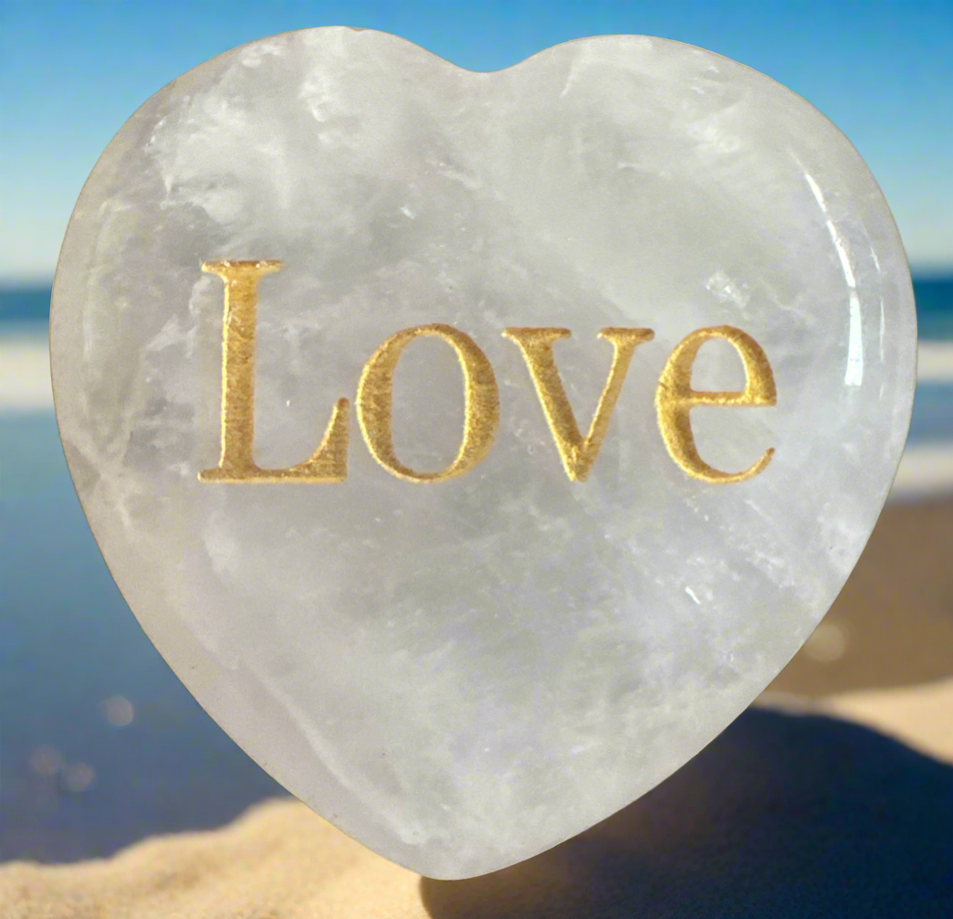 Heart-shaped white quartz stone with the word "Love" in gold lettering, resting on sand with the ocean in the background.
