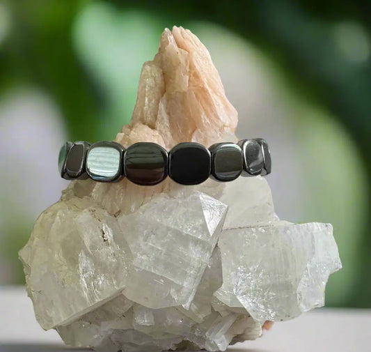 Bracelet made of shiny, black square beads, resting on two minerals with a blurred green background.