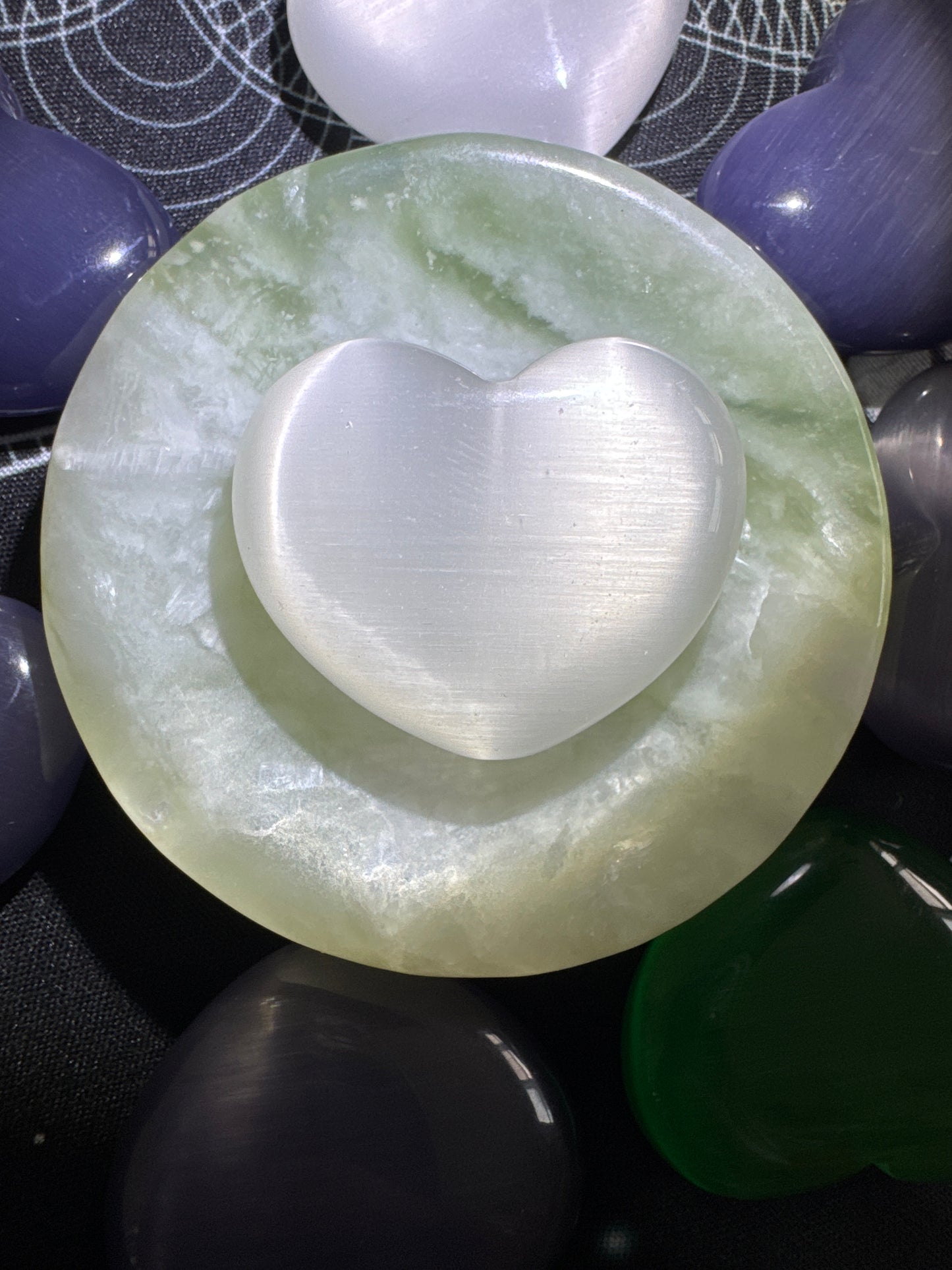 Overhead shot of a white heart-shaped selenite crystal on a green round marble disc, surrounded by polished heart-shaped crystals in various colors including purple, gray, green, and white, all on a black surface with a textured geometric pattern.