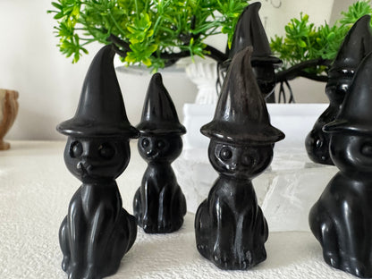 Group of black cat figurines wearing pointed witch hats, displayed on a white textured surface, with a green plant in the background.