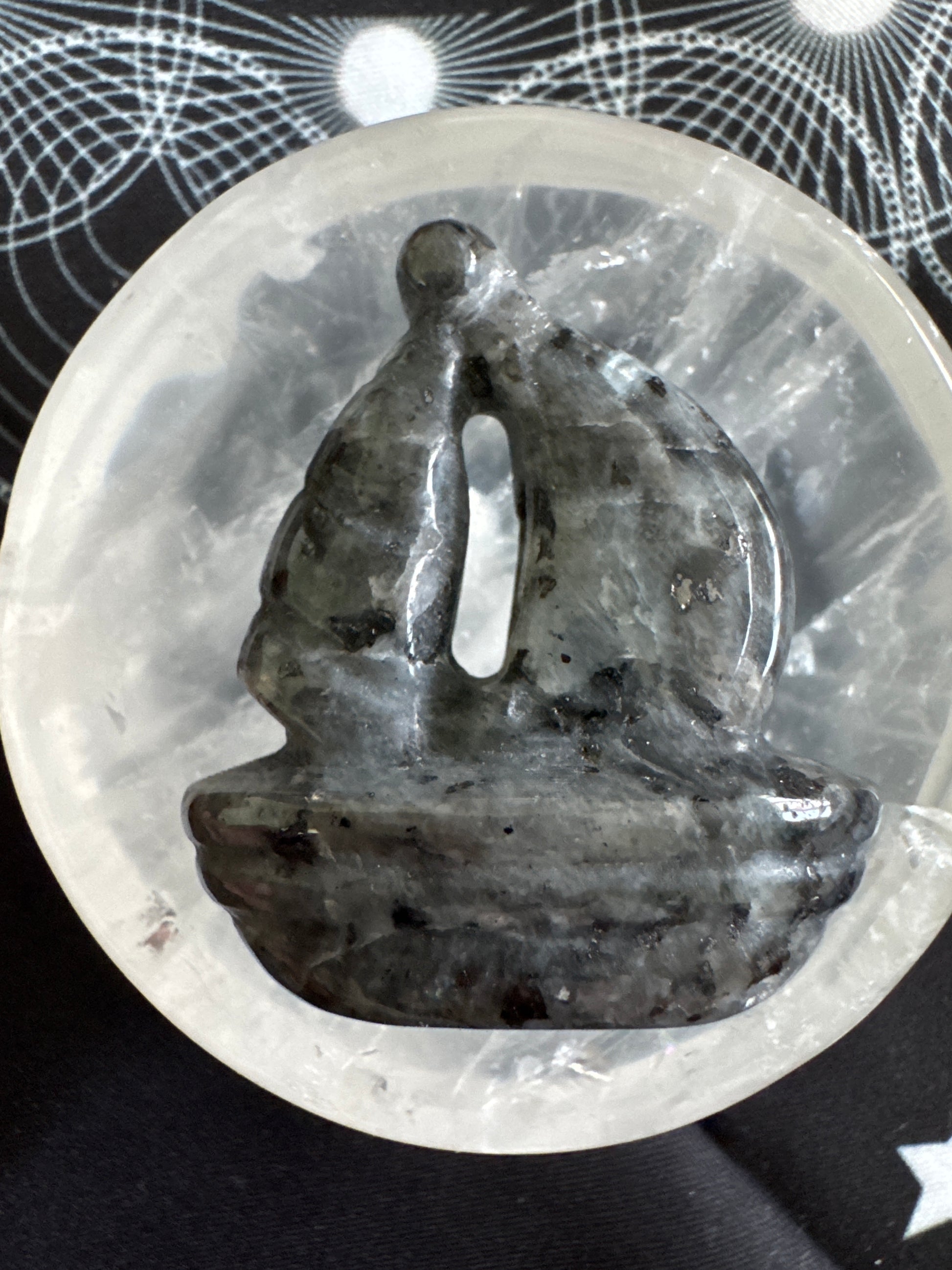 Close-up of a gray stone sculpture with a vertical slit, nestled in a clear quartz bowl on a patterned fabric.