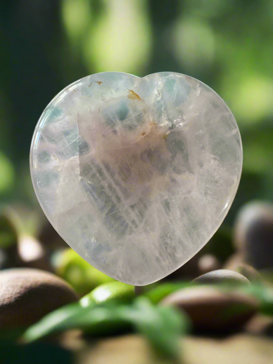 Translucent, heart-shaped crystal with natural imperfections, set against a blurred background of rocks and greenery.